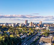 A wide aerial view of a city skyline with skyscrapers, highways, and trees under a blue sky and scattered clouds.