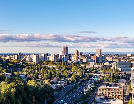 A wide aerial view of a city skyline with skyscrapers, highways, and trees under a blue sky and scattered clouds.