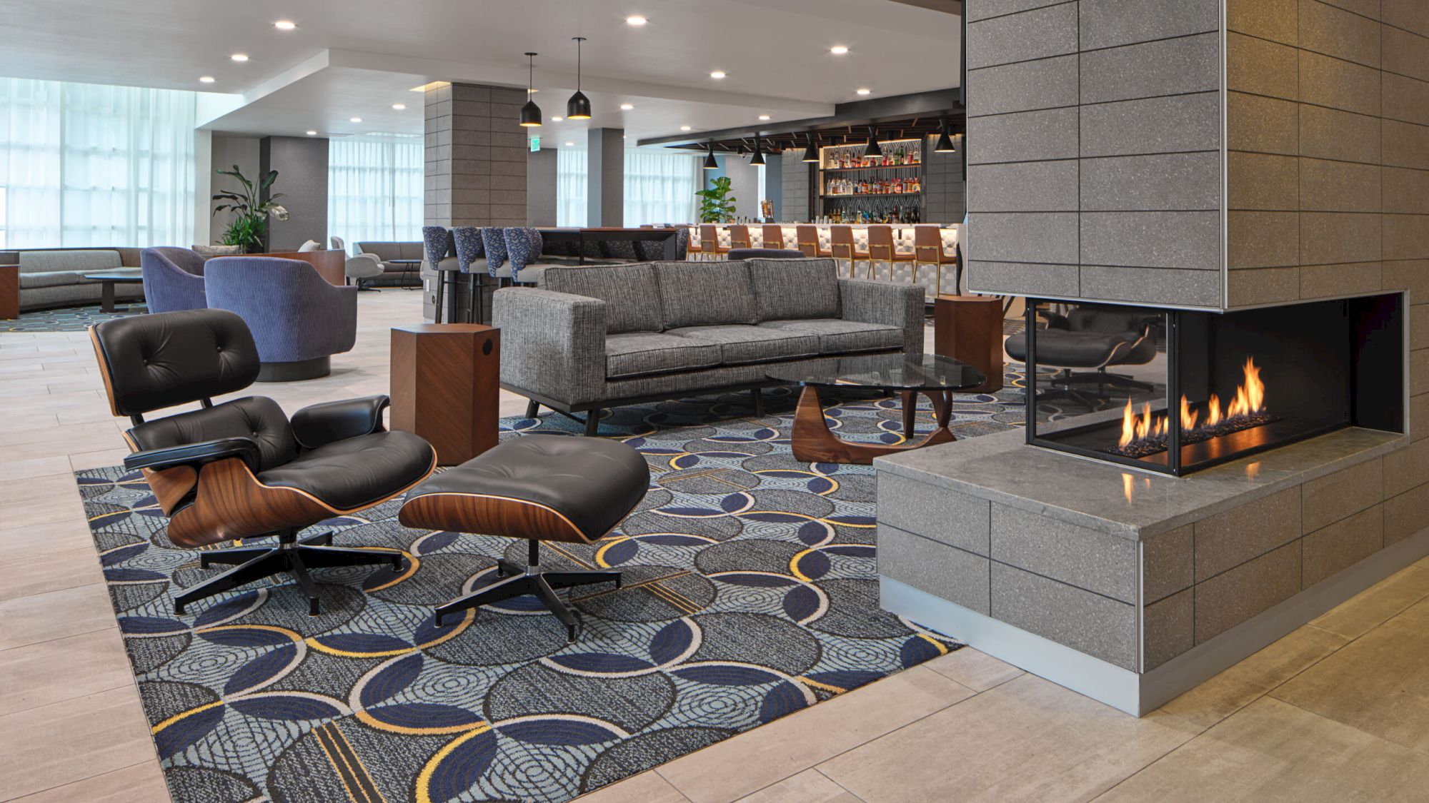 A modern lounge with gray sofas, a patterned rug, a fireplace, and a bar area in the background.