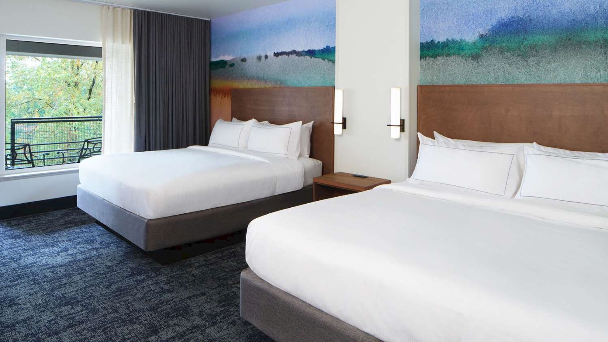 The image shows a hotel room with two large beds, a colorful wall mural, a window with a view, and modern lighting fixtures.