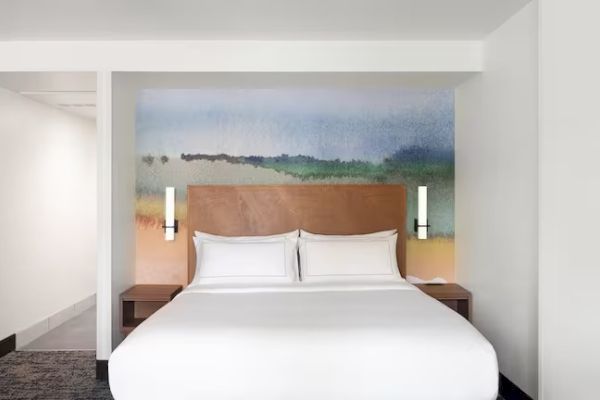 A modern hotel room features a large bed with white linens, two mounted lights, and a colorful mural on the wall behind it.