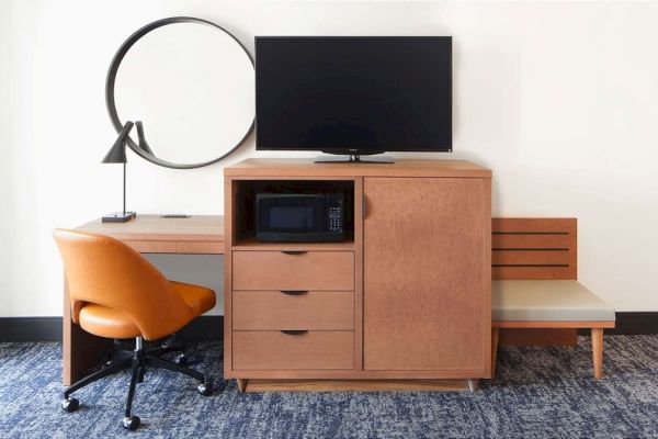 A room setup with a desk, chair, lamp, microwave, drawers, TV, and seating bench.