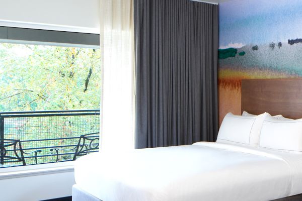 The image shows a bright hotel room with a bed, colorful wall art, large window with curtains, and a view of trees.