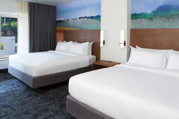 The image shows a hotel room with two large beds, modern decor, and a mural on the wall. A window provides natural light.