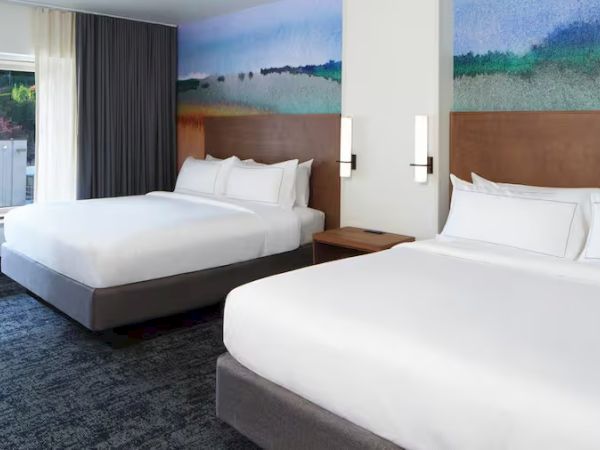 The image shows a hotel room with two large beds, modern decor, and a mural on the wall. A window provides natural light.