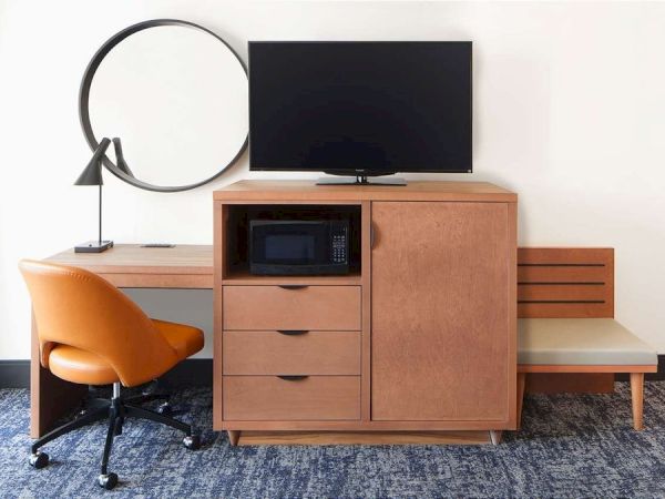 The image shows a modern hotel room setup with a desk, orange chair, round mirror, TV, cabinet, and microwave on a blue carpet.