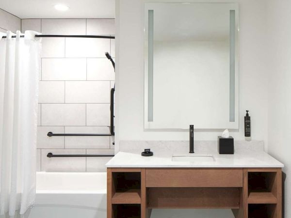 The image shows a modern bathroom with a bathtub, white shower curtain, vanity with mirror, sink, and wooden shelves, all in a minimal design.