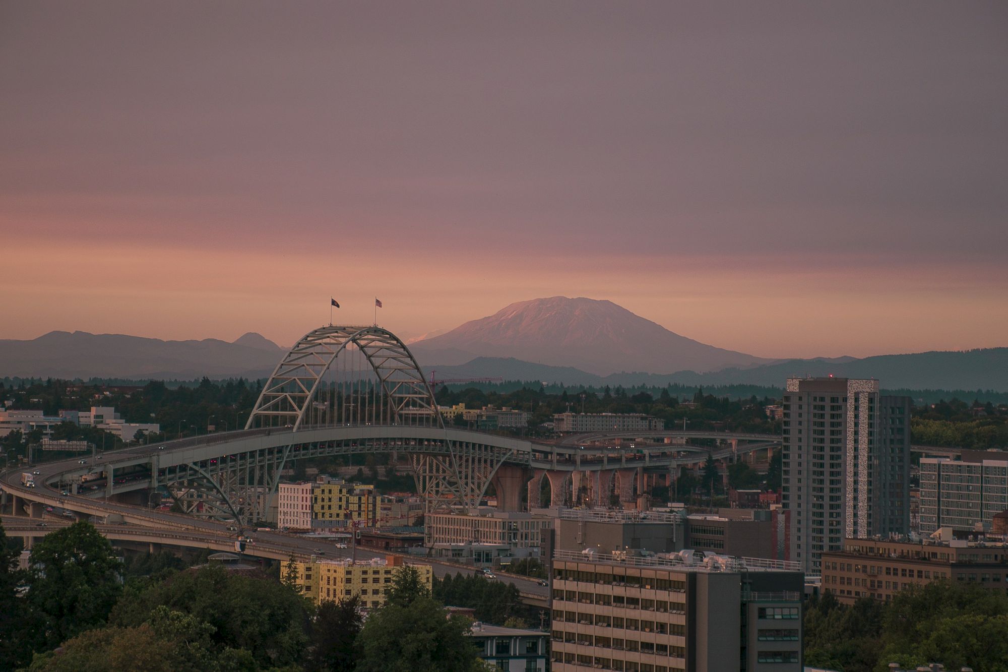 A cityscape at sunset featuring a large arch bridge, buildings, and a distant mountain under a colorful sky.
