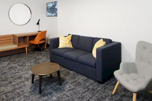 A modern room with a gray sofa, two yellow pillows, a round table, an armchair, a desk, and a circular wall mirror.