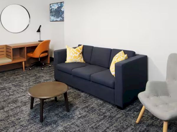 A modern room with a gray sofa, two yellow pillows, a round table, an armchair, a desk, and a circular wall mirror.