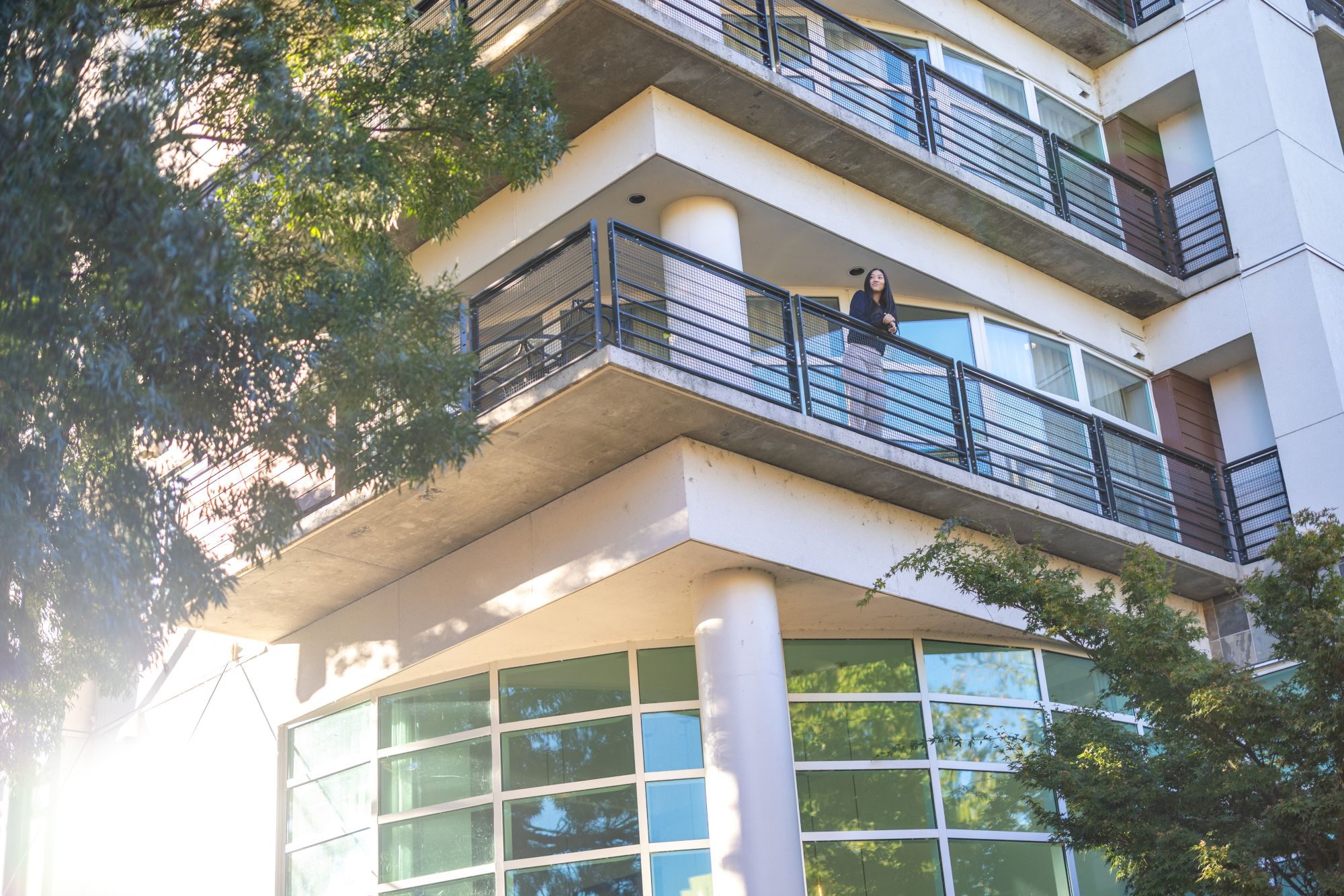 A modern multi-story building with glass windows and balconies; a person leans on the railing of the top floor balcony.