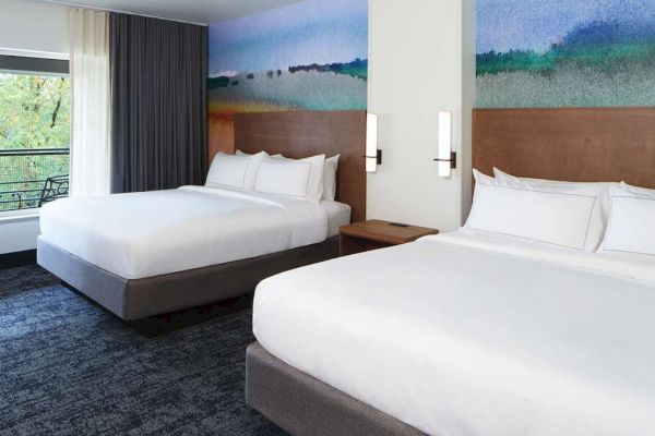 The image shows a hotel room with two large beds, a colorful wall mural, a window with a view, and modern lighting fixtures.