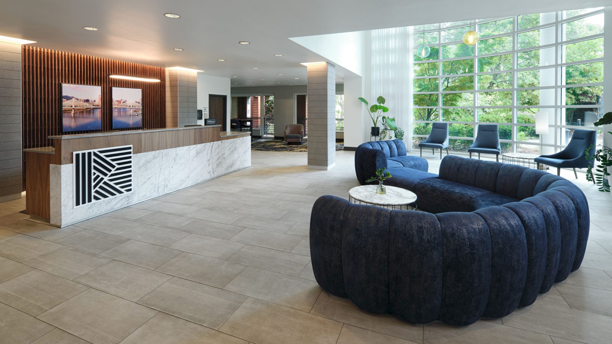The image shows a modern lobby with a marble reception desk, dark blue sofa, chairs, large windows, and greenery visible outside.