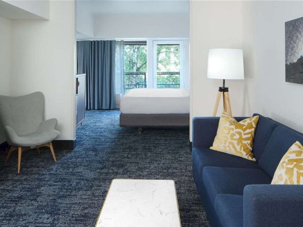 A cozy hotel suite features a fireplace, armchair, sofa with yellow pillows, and a bed by large windows with blue curtains.