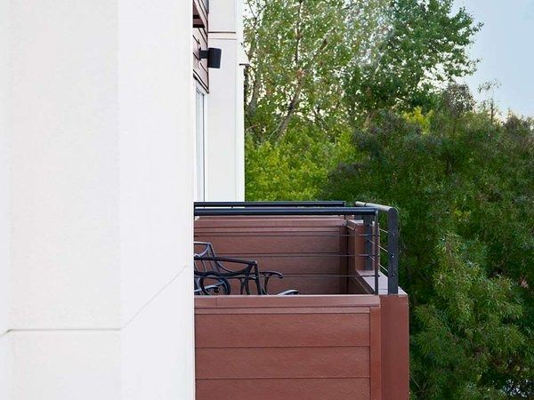 A building's side view shows balconies with chairs, set against lush green trees and a clear sky.