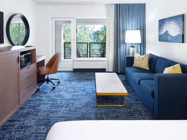 A modern hotel room features a bed, blue sofa, wooden desk with a chair, large TV, lamp, art on the wall, and a view outside the window.