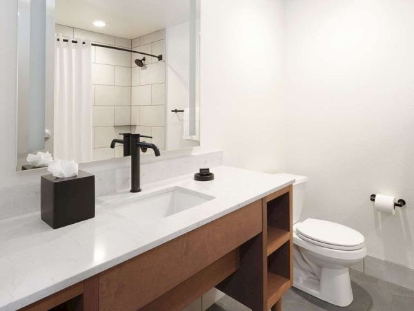 This image shows a modern bathroom with a sink, mirror, black faucet, white countertop, toilet, and tiled shower with curtain.