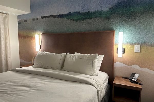 A hotel room with a bed, colorful headboard art, two wall lights, a nightstand with a phone, and a wall switch visible.
