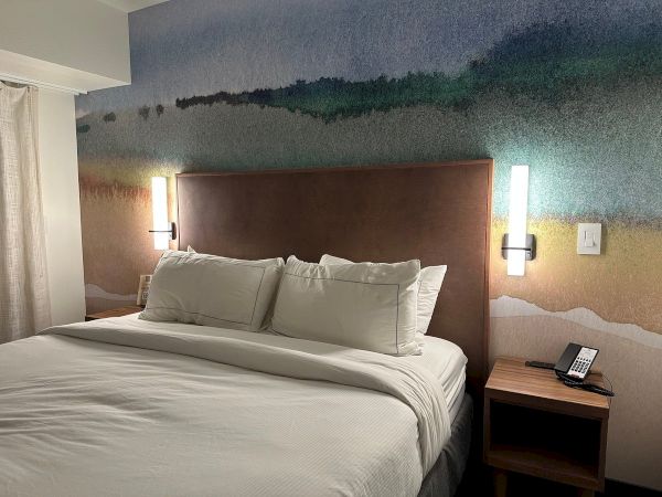 A hotel room with a bed, colorful headboard art, two wall lights, a nightstand with a phone, and a wall switch visible.