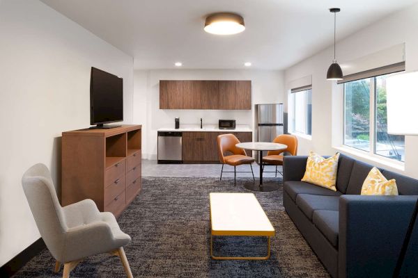 A modern living room with a TV, sofa, table, chairs, and a kitchenette with appliances. It has neutral tones and bright accents against the sentence.