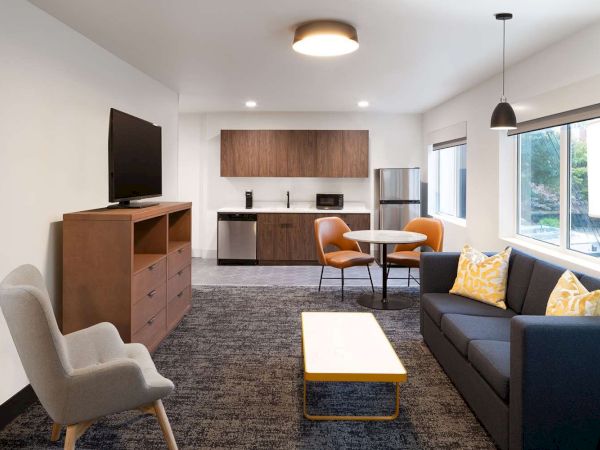 A modern living room with a TV, sofa, table, chairs, and a kitchenette with appliances. It has neutral tones and bright accents against the sentence.