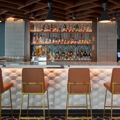 A stylish bar with geometric decor, high chairs, and a well-stocked shelf of liquor bottles under soft lighting.