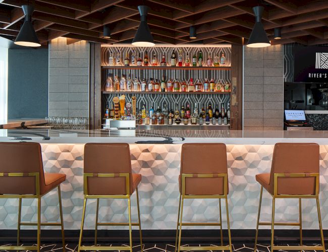 A stylish bar with geometric decor, high chairs, and a well-stocked shelf of liquor bottles under soft lighting.