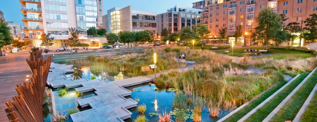 An urban park with water features, plants, and seating areas, surrounded by modern buildings at dusk, with a few people relaxing nearby.