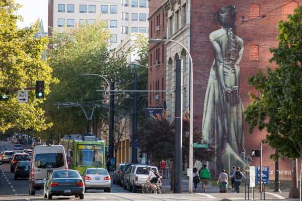 A bustling street scene with cars, a tram, and pedestrians. A large mural of a woman on a brick building adds artistic flair to the view.