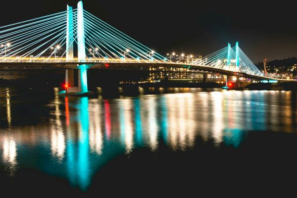 A cable-stayed bridge lit up at night, reflecting beautifully on the calm river water below.