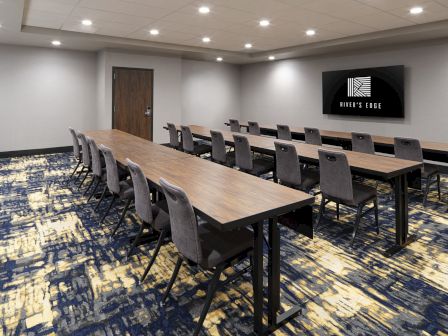 A modern conference room with long tables, chairs, and a wall-mounted screen, featuring a patterned carpet and neutral walls.