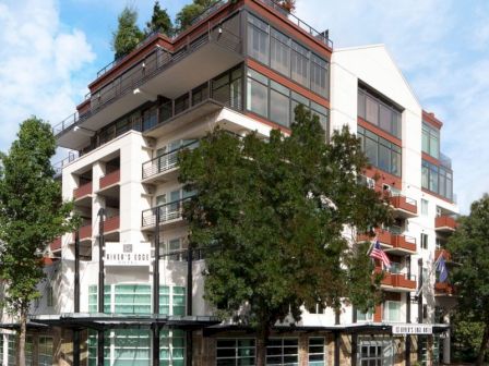 The image shows a corner view of a modern multi-story apartment building with balconies and greenery on the rooftop.