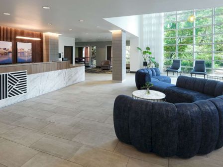 A modern lobby with a marble reception desk, curved blue sofa, and large windows letting in natural light. Contemporary artwork adorns the wall.