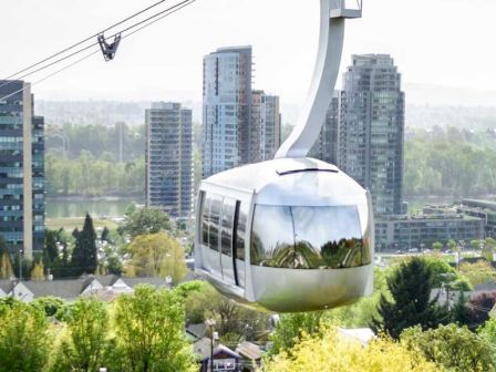 A modern cable car travels above a cityscape with tall buildings and lush greenery in the background, reflecting an urban and natural mix.