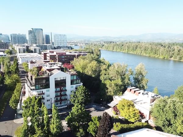 An aerial view of a riverside cityscape with modern buildings, lush greenery, and a river under a clear blue sky.