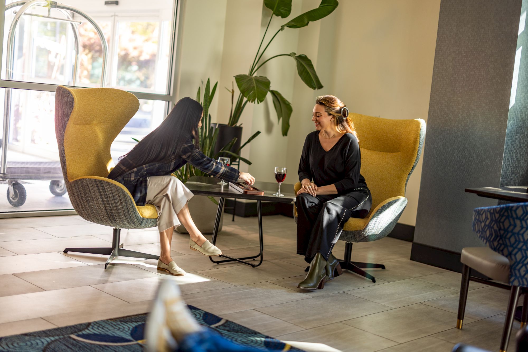 Two people sit in modern chairs, legs crossed, chatting over drinks in a bright lounge with plants and large windows.
