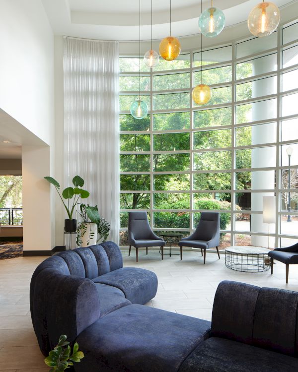 A modern lounge with a curved blue sofa, large windows, hanging lights, and plants, creating a serene atmosphere with natural light.