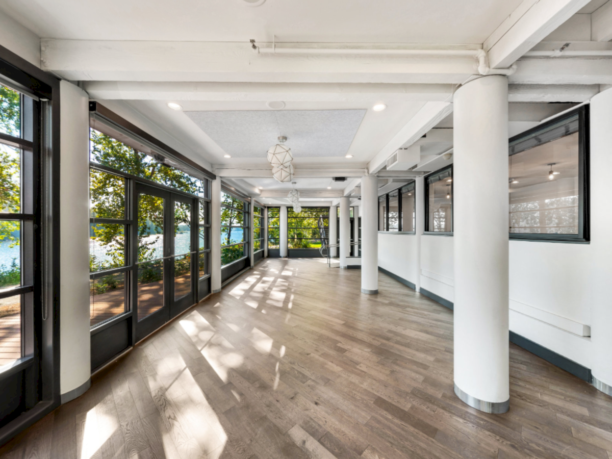 A bright modern corridor with tall windows, white columns, wooden floors, and overhead lights with trees outside.