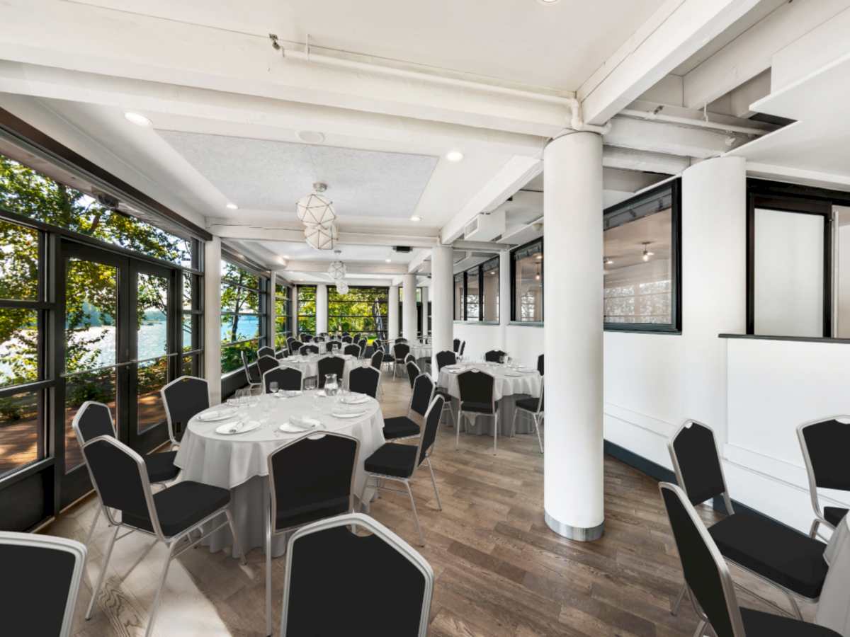 A bright banquet hall with set tables and chairs, hanging lights, and large windows revealing trees and a lake outside.