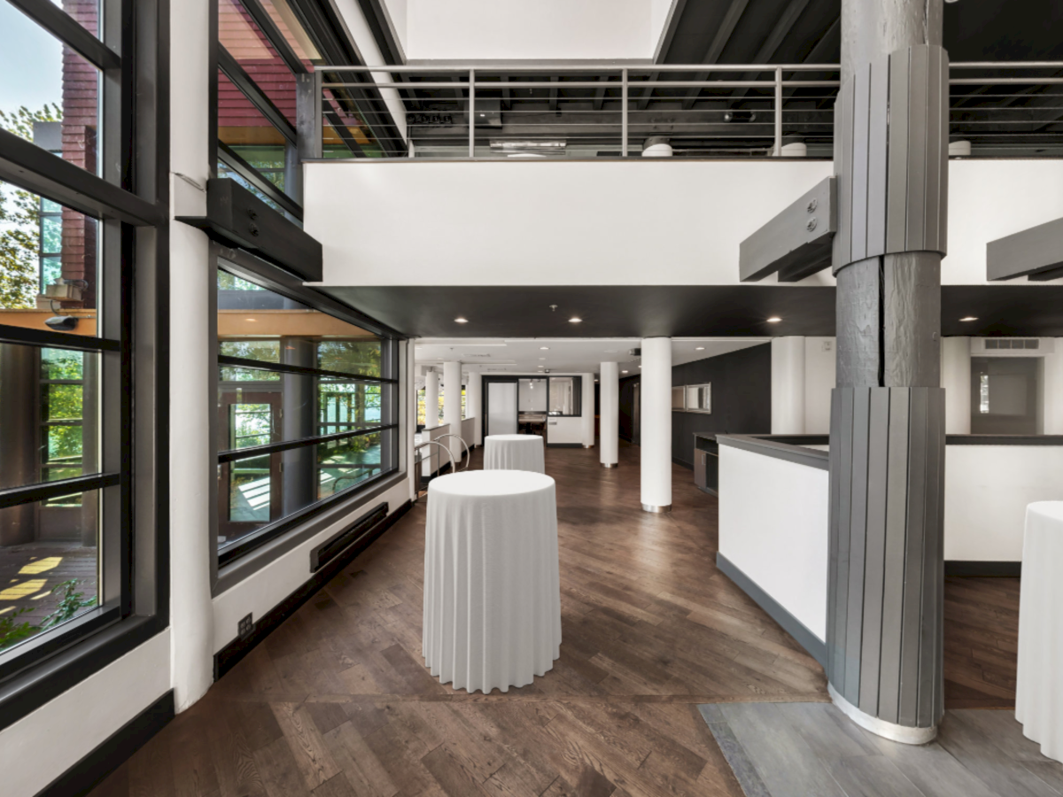 A modern indoor event space with large windows, white-covered tables, and sleek dark ceiling beams.