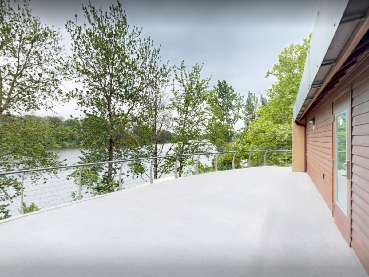 A lakeside deck beside a house, with a metal railing and green trees under a cloudy sky, offering a wide water view.