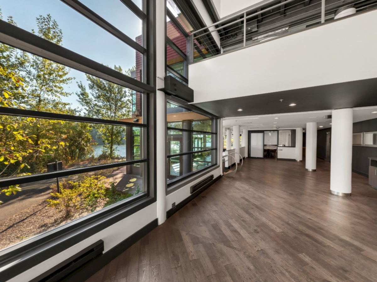 A modern indoor corridor with large floor-to-ceiling windows, white columns, and warm wood flooring.