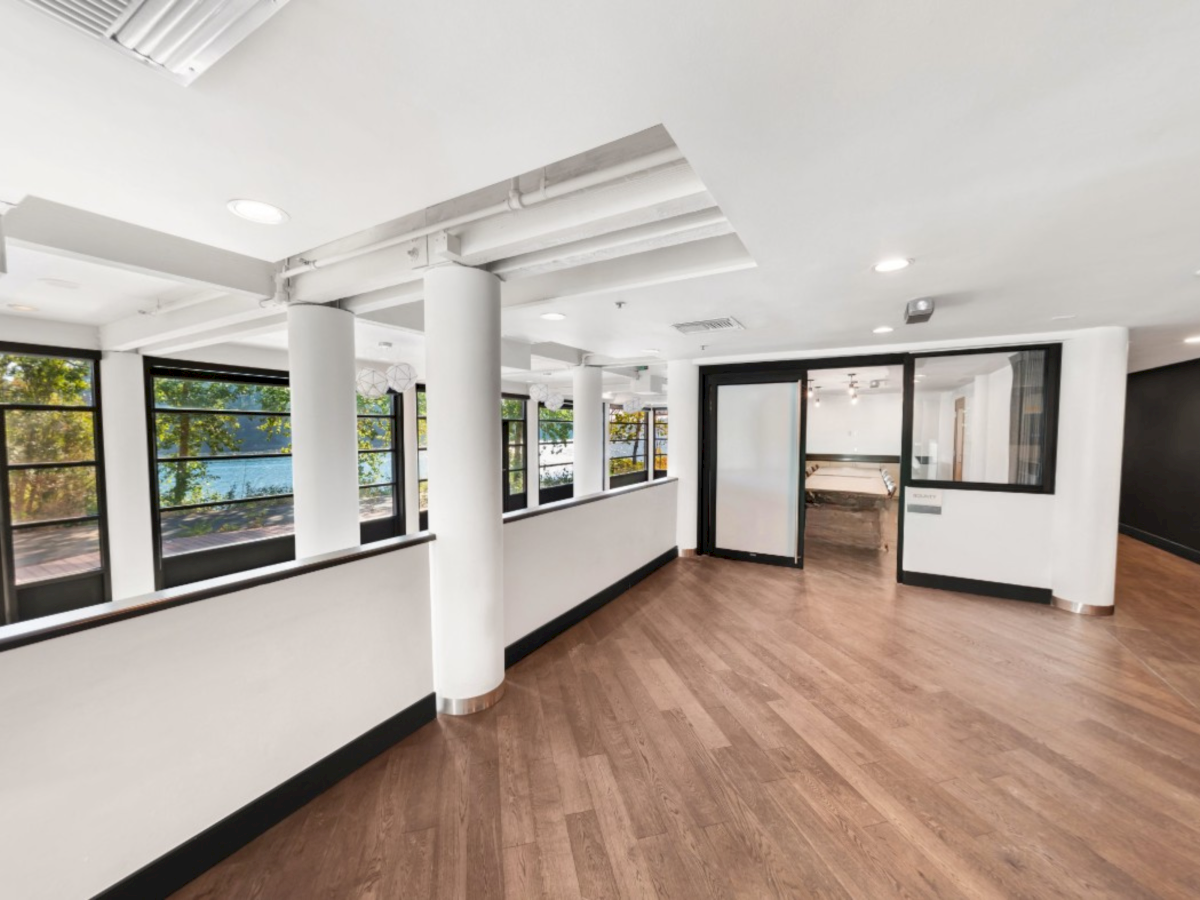 Bright modern office lobby with wood floors, white columns, black-framed glass walls, and skylight-style ceiling lights.