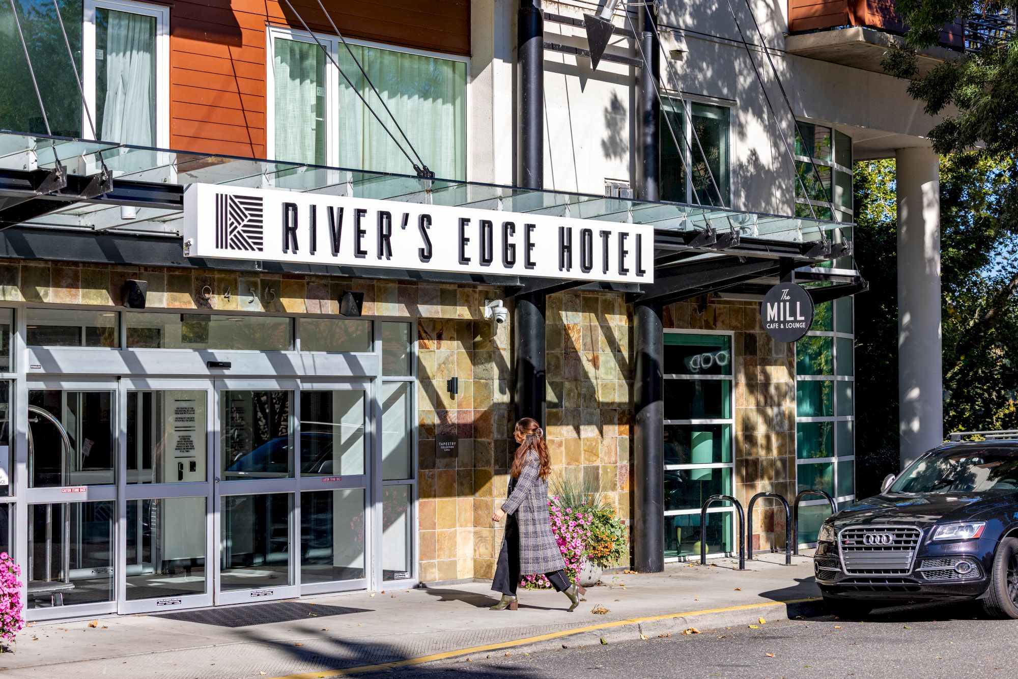 A street view of River’s Edge Hotel with a glass storefront, a couple walking by, and a parked car by the entrance.