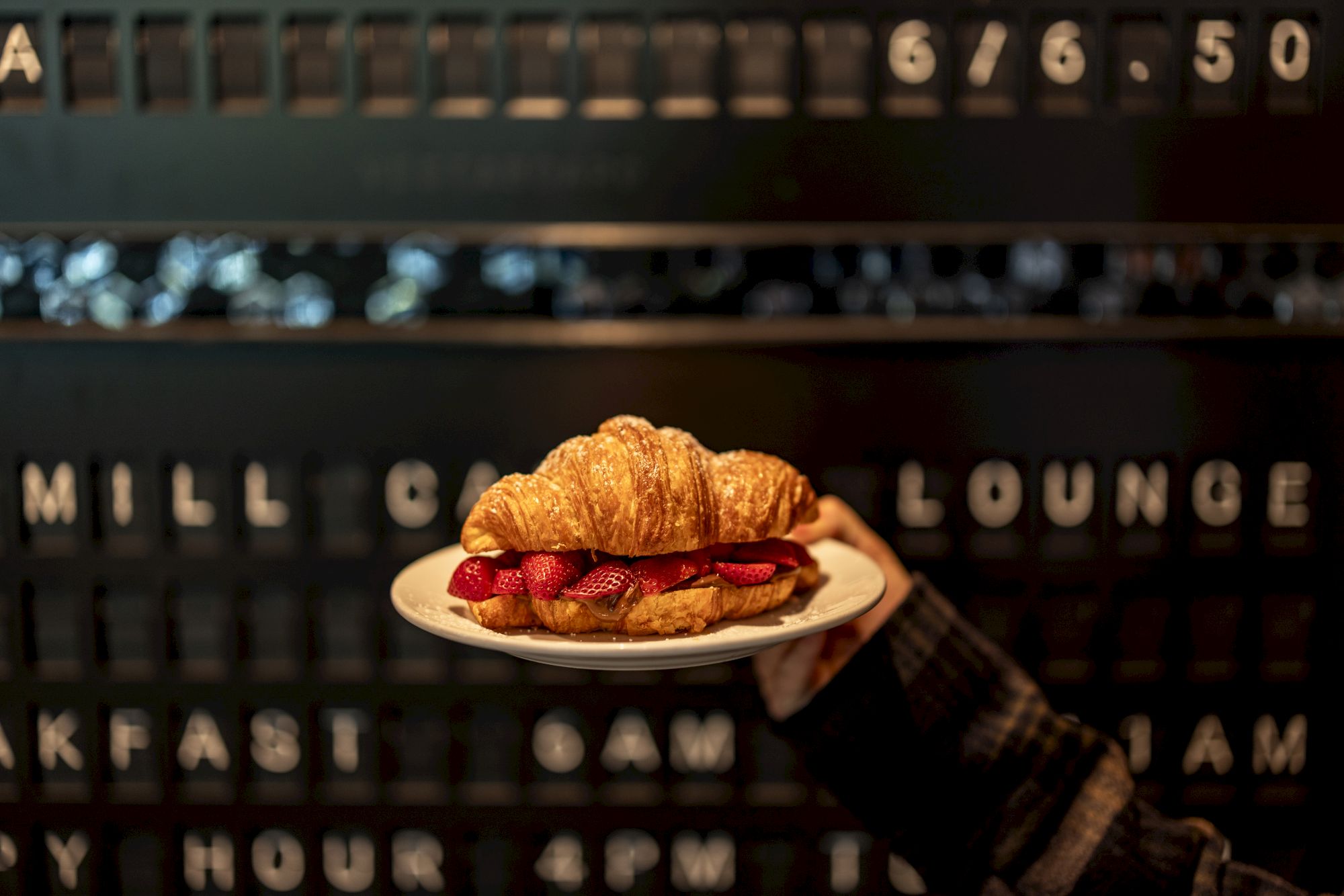 A hand holds a plate with a croissant sandwich against a dark, retro menu board backdrop.