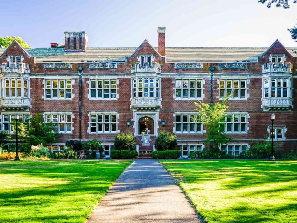 A brick collegiate building with large white-trim windows, a central arched doorway, and a green lawn leading to a paved path&mdash;classic campus vibes.