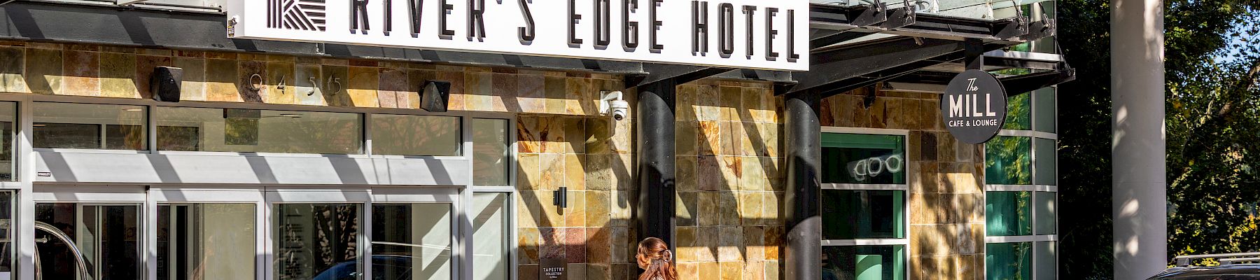 A hotel entrance labeled River’s Edge Hotel and The Mill Cafe & Lounge, with a woman walking past flowers and a parked Audi car.