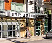 A hotel entrance labeled River’s Edge Hotel and The Mill Cafe & Lounge, with a woman walking past flowers and a parked Audi car.