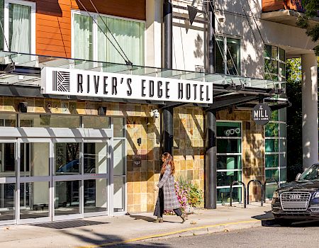 A hotel entrance labeled River’s Edge Hotel and The Mill Cafe & Lounge, with a woman walking past flowers and a parked Audi car.