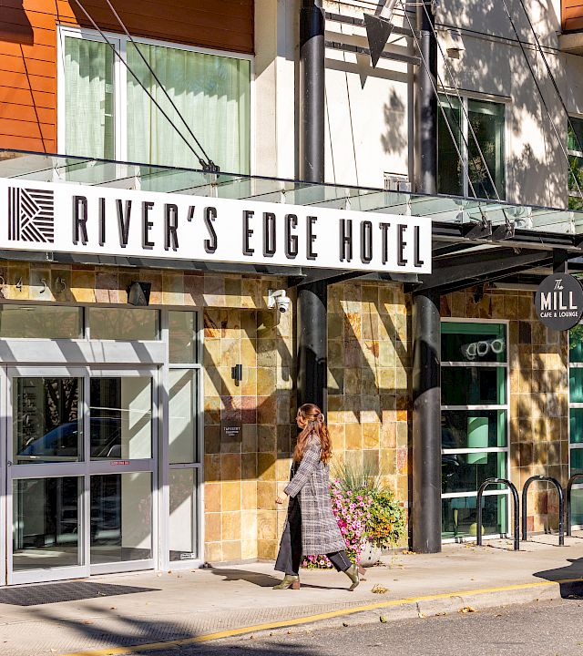 A hotel entrance labeled River’s Edge Hotel and The Mill Cafe & Lounge, with a woman walking past flowers and a parked Audi car.